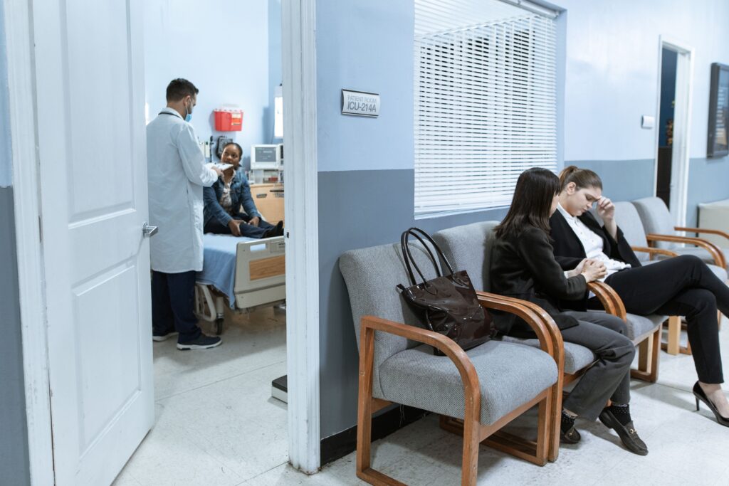 Patients waiting in a hospital emergency department while a doctor examines a patient in a nearby treatment room, representing ED overcrowding and patient wait times.
