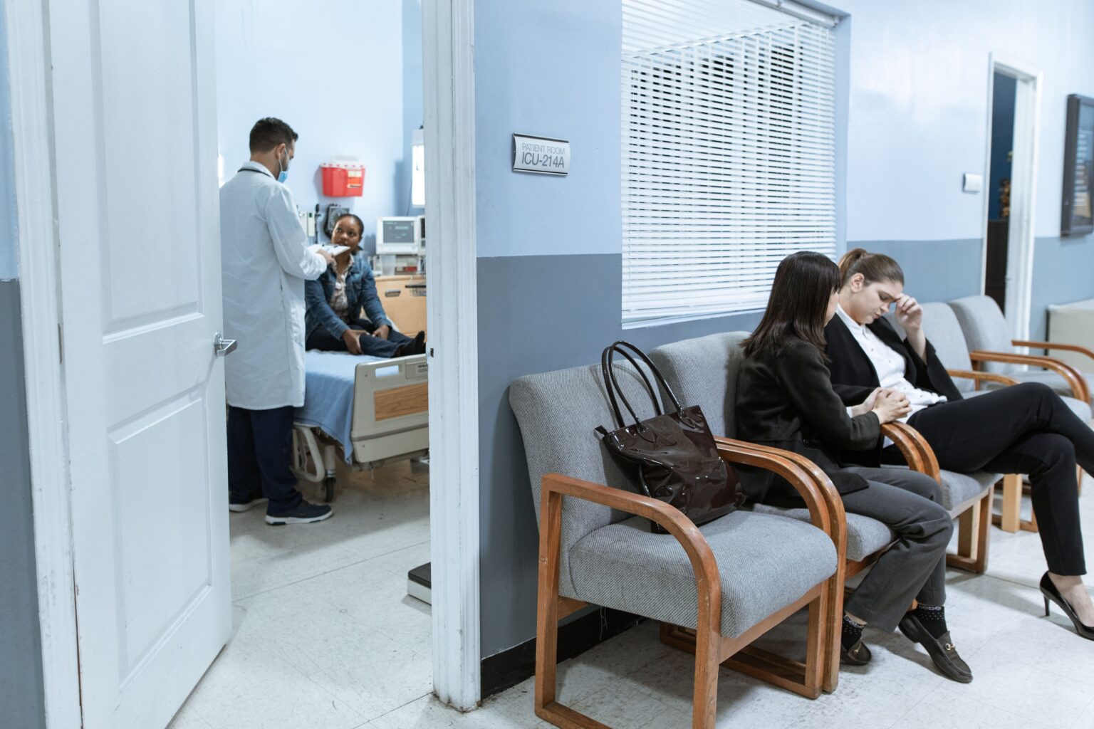 Patients waiting in a hospital emergency department while a doctor examines a patient in a nearby treatment room, representing ED overcrowding and patient wait times.